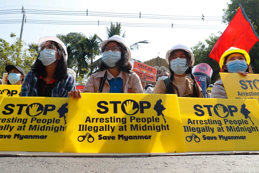 Four young women in face masks hold yellow signs reading: "Stop arresting people illegally at midnight save Myanmar".