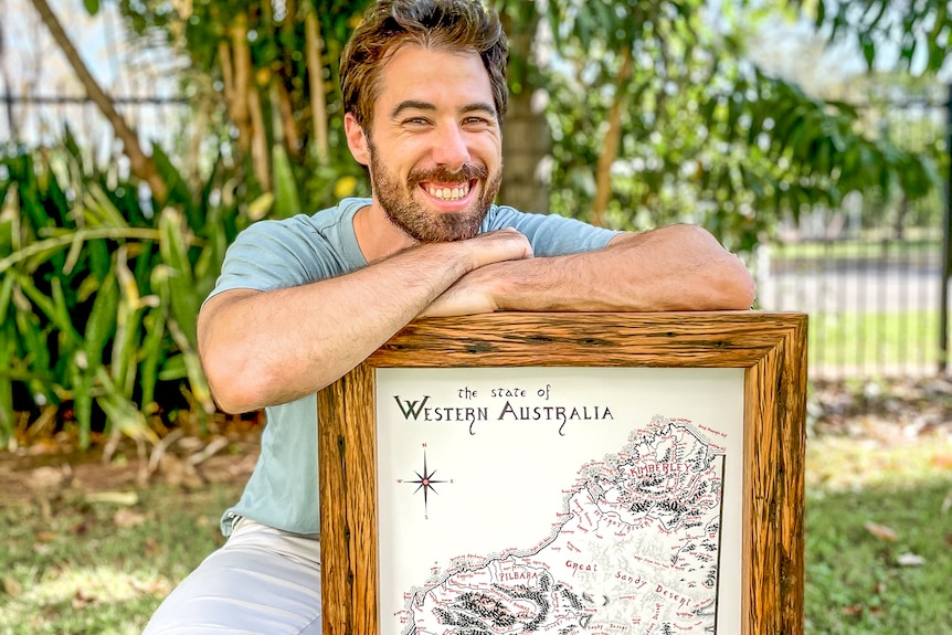 A man with brown hair and a beard crouches down by a framed hand-drawn map of Western Australia smiling.