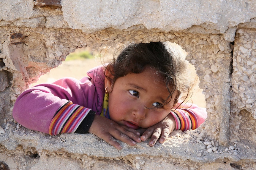 A little girl stares through a hole in a stone fence.