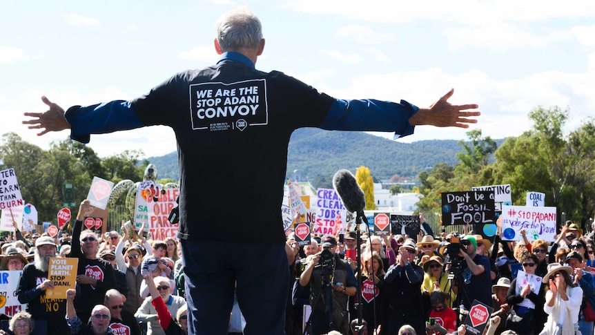 Bob Brown standing before the rally with his arms spread widely with a "We are the stop Adani convoy" shirt.