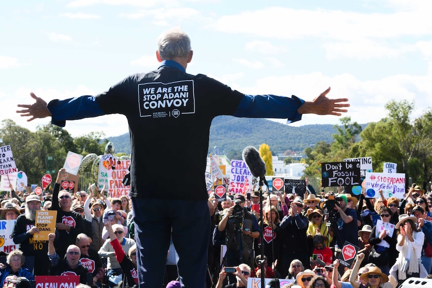 Bob Brown standing before the rally with his arms spread widely with a "We are the stop Adani convoy" shirt.