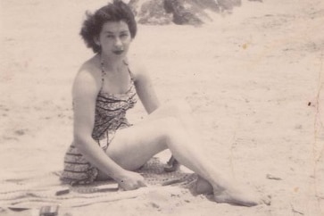 Black and white grainy photo of young woman in full-piece bathing suit sitting on the sand.