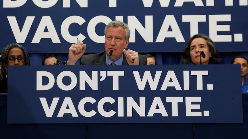 A woman and a man sit behind microphones at a desk with a banner reading "Don't wait. Vaccinate".