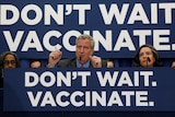 A woman and a man sit behind microphones at a desk with a banner reading "Don't wait. Vaccinate".