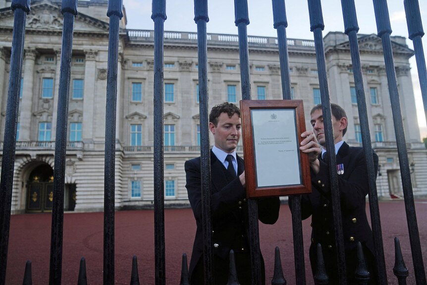 Two men dressed in dark suits and wearing a tie put up a framed document on a fence.