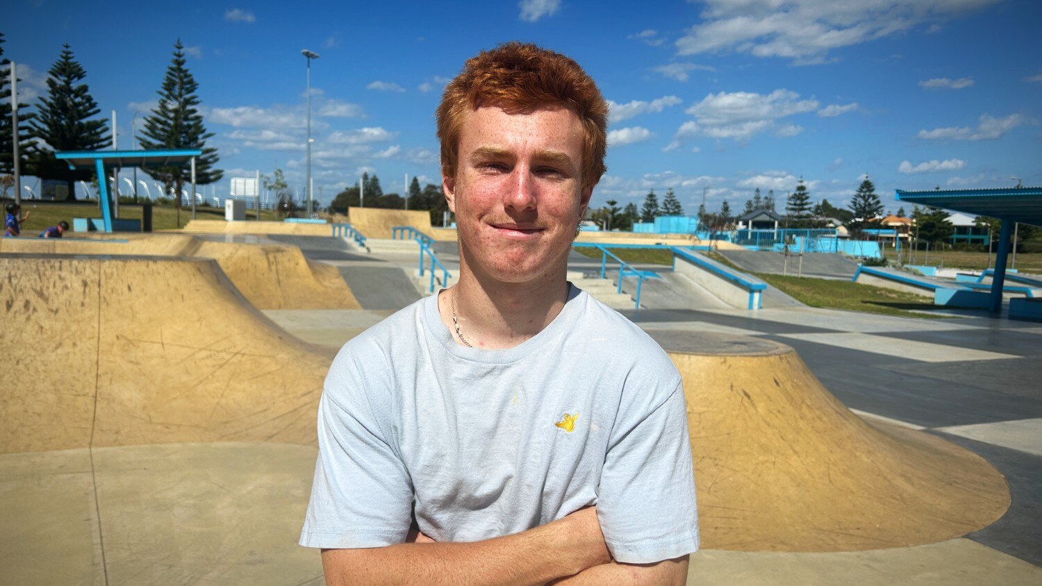 A young man with red hair wearing a black hoodie holding a skateboard at night near a skate park