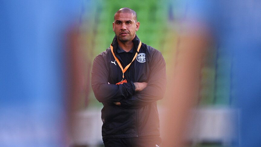 Patrick Kisnorbo watches training with two players standing blurred in the foreground