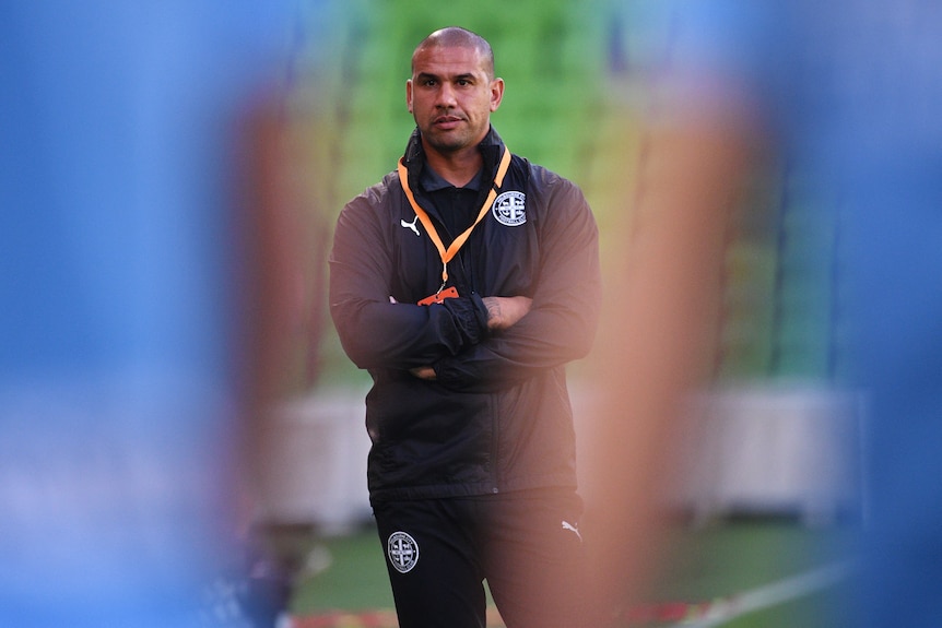 Patrick Kisnorbo watches training with two players standing blurred in the foreground