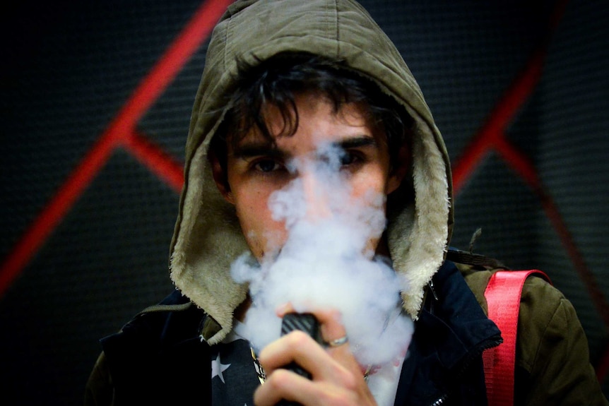 a young man wearing a hooded jumper sucks on an e-cigarette blowing out smoke which conceals his face.
