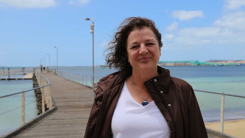 A woman with curly brown hair, wearing a white t-shirt stands in front of a seaside jetty.