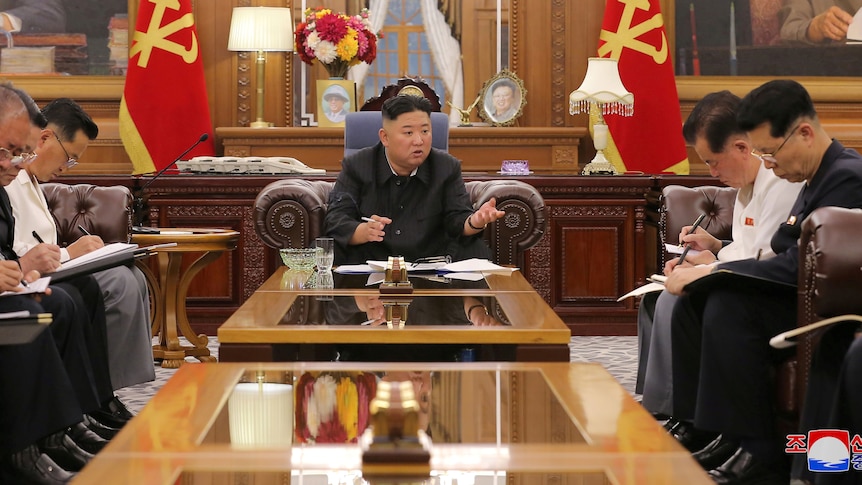 Kim Jong Un sitting at a table, speaking to assembled men 