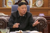 Kim Jong Un sitting at a table, speaking to assembled men 