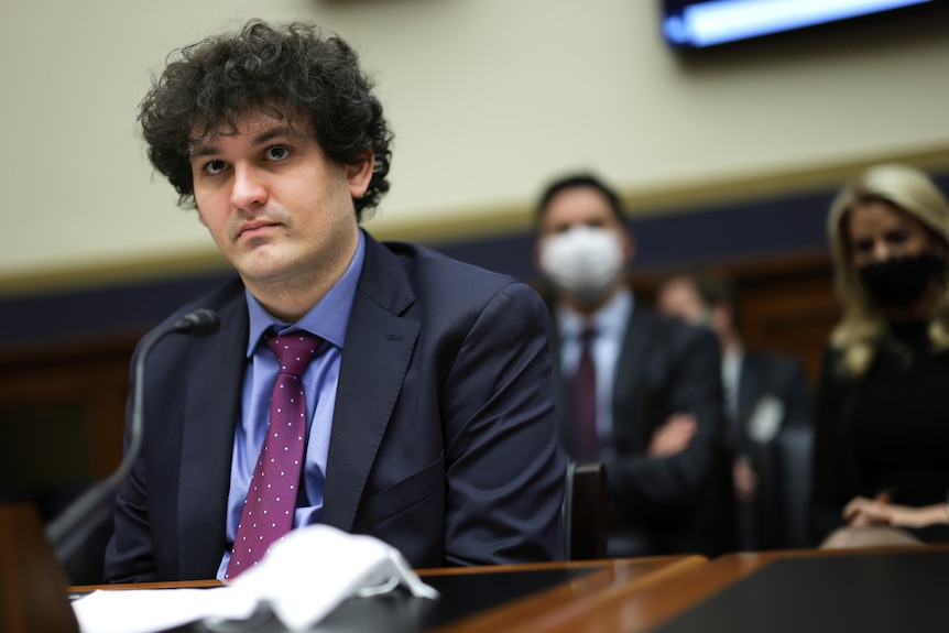 Sam Bankman Fried in a suit sits before a microphone on a desk 