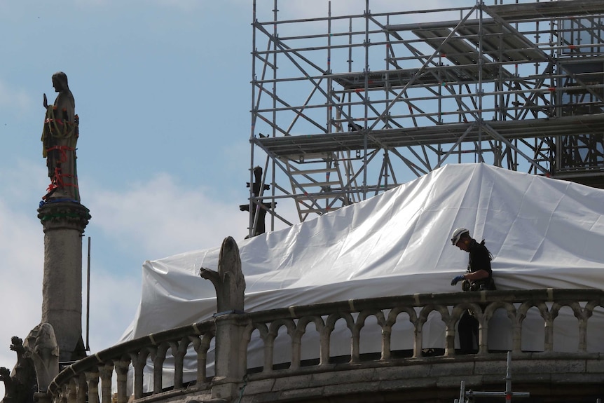 Looking from below, you see a worker on Notre Dame's neoclassical roof with scaffolding and and white tarp behind him.