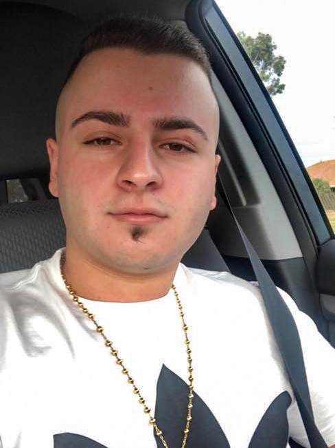 Savio Mansour, wearing a gold chain and white Adidas jumper, takes a selfie in a car.
