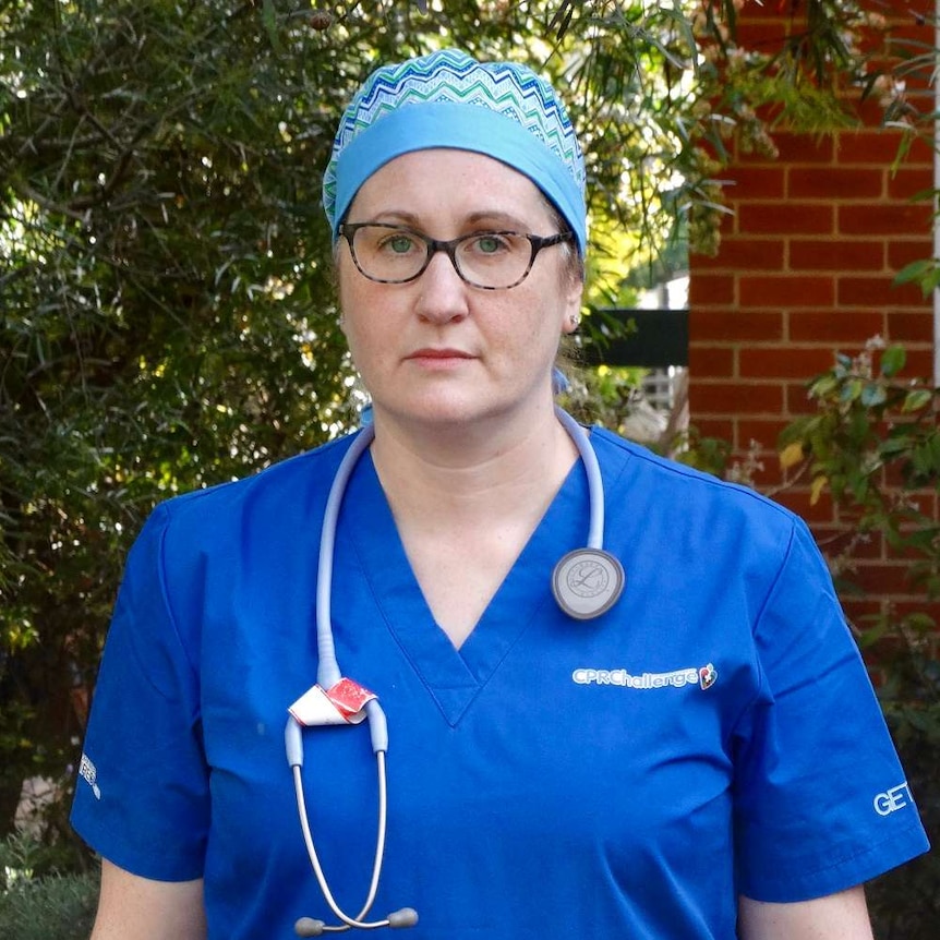 Claire Wilkin-Marshall dressed in blue scrubs and glasses stands outside with a serious expression.