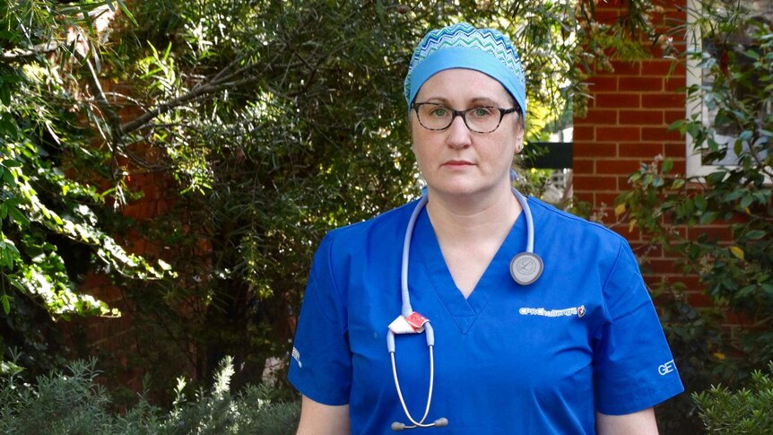 Claire Wilkin-Marshall dressed in blue scrubs and glasses stands outside with a serious expression.