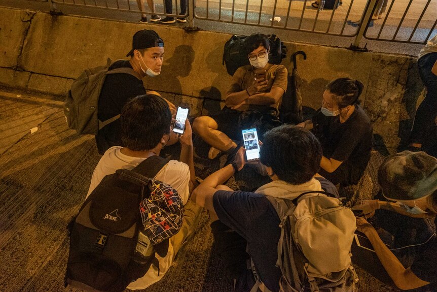 A group of young people, some wearing backpacks and face masks, sit in a circle and look at their smartphones.
