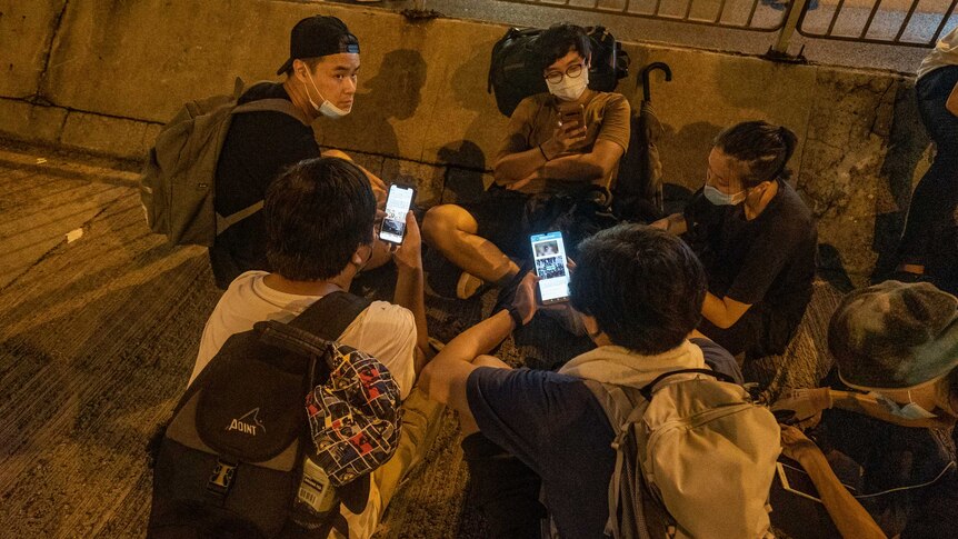 A group of young people, some wearing backpacks and face masks, sit in a circle and look at their smartphones.