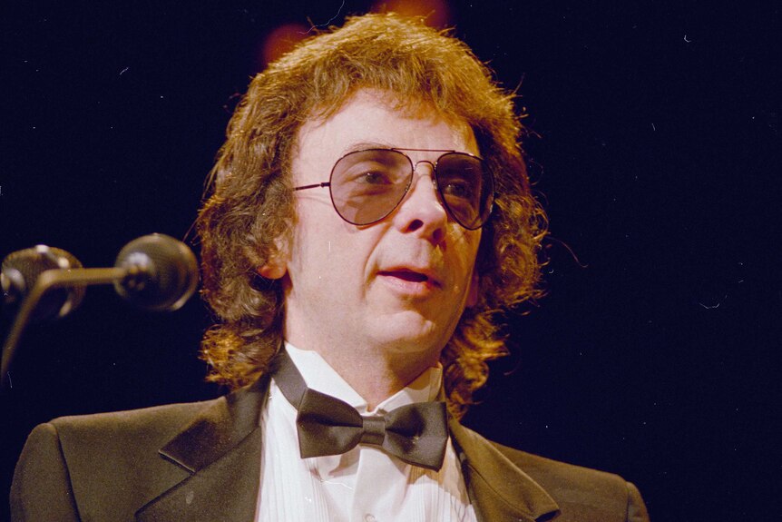 A bespectacled man with shaggy hair wears a tuxedo as he speaks into a microphone on a stage.