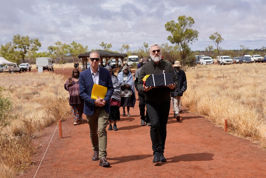 Two men leading a group of Indigenous people. The man on the right carries a box of remains.