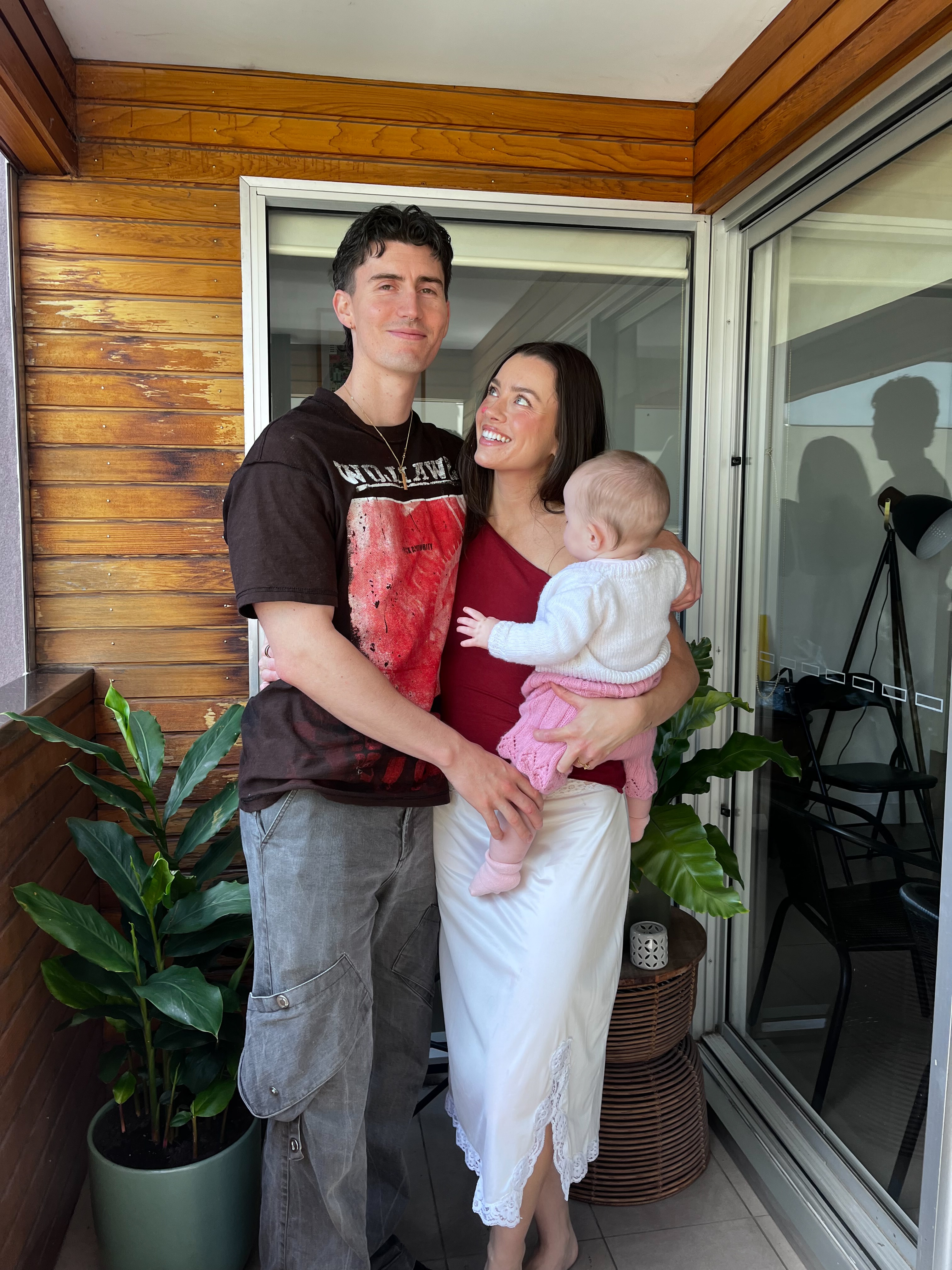 Madeleine Edwards holds her baby Carmine on her hip and smiles with her partner Matt on a balcony.