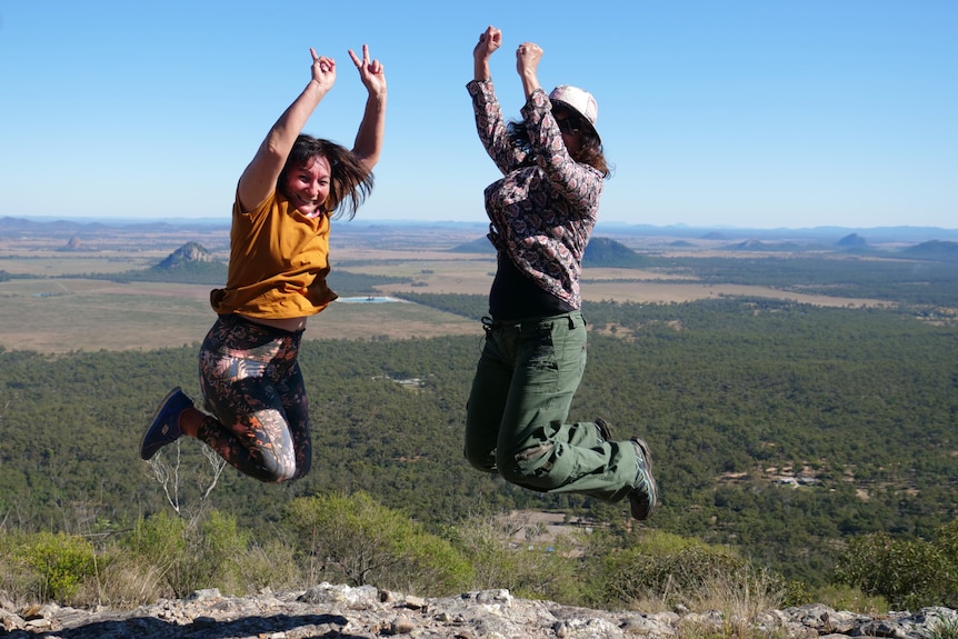 Two ladies with their hands in the air and knees up, both hovering, mountains, trees and blue sky in the background.