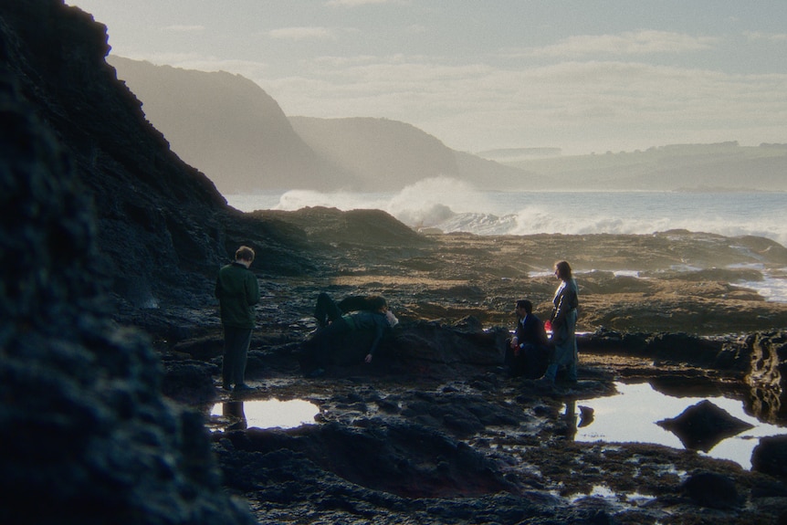 On a rocky beach, three young people look on at a woman leans into a young man prostrate on a rock