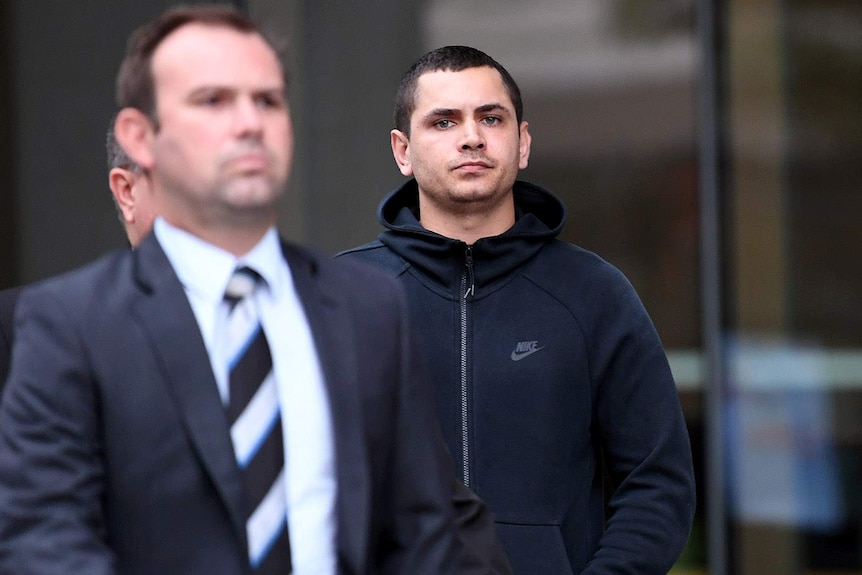 A man in a black hoodie stares at the camera, standing behind another man in a suit, slightly blurred