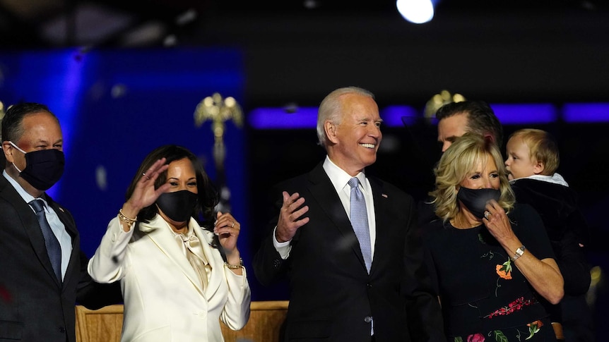 Joe Biden smiles, standing in front of Kamala Harris and Jill Biden as they celebrate his presidential election win