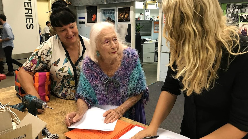 A woman fills out paperwork as two other women stand either side of her.