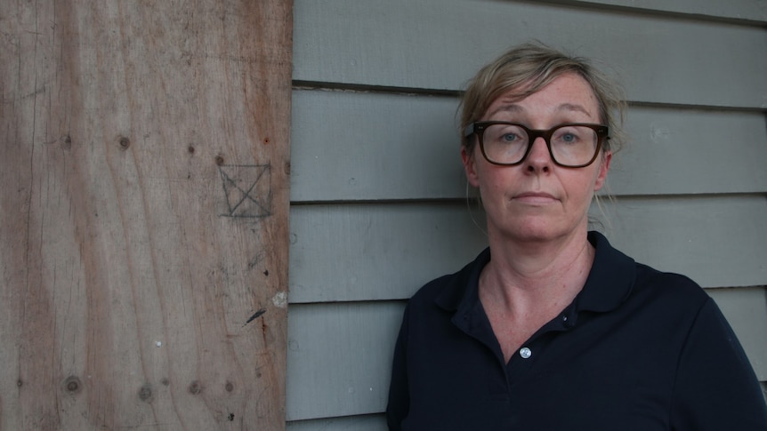 Woman stares at camera with glasses standing infront of partially boarded home