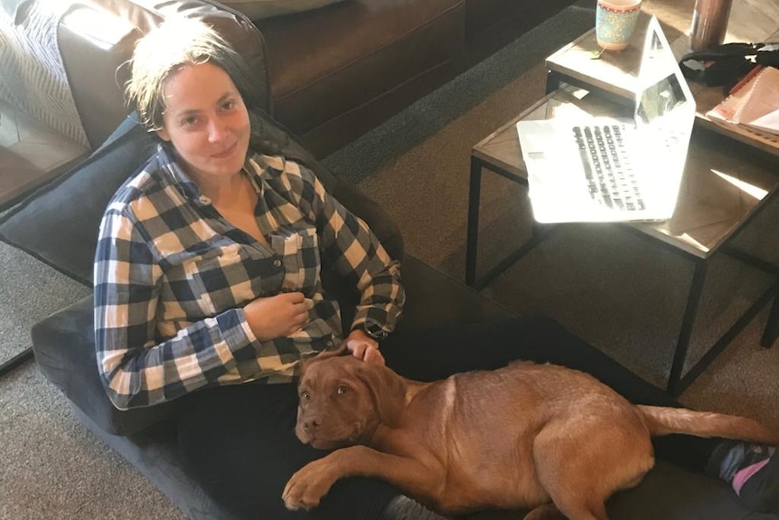 A woman in a checked shirt laying on the floor with a reddish-brown dog