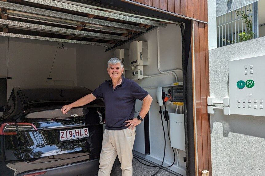 Hombre con cabello canoso y polo azul marino parado en su garaje, apoyado en su vehículo eléctrico