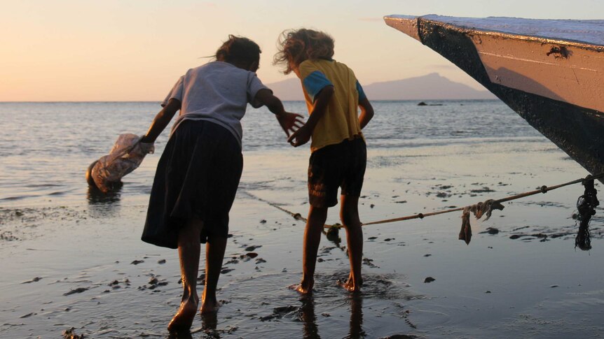 Two Timorese children search for shellfish on a beach, with the island of Atauro in the background.