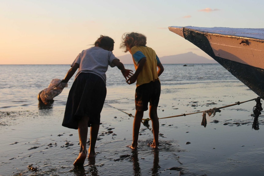 Two Timorese children search for shellfish on a beach, with the island of Atauro in the background.