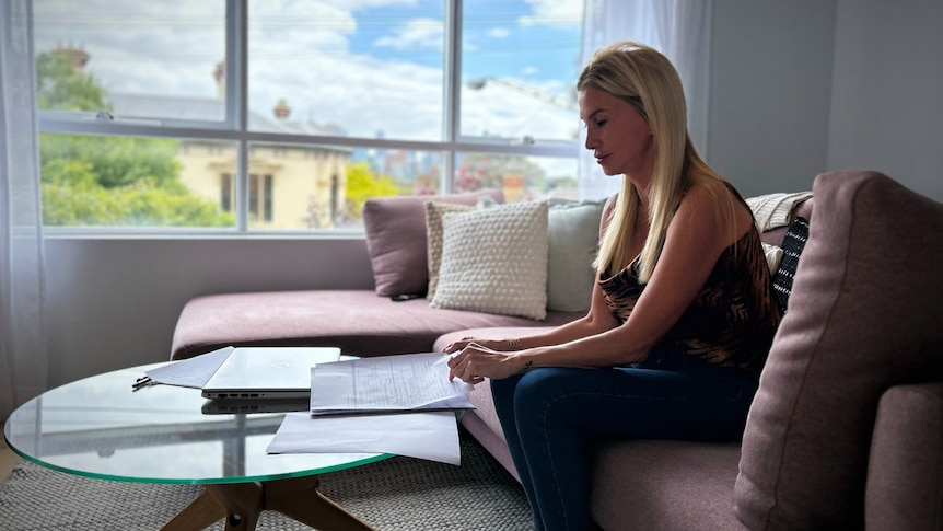 A woman with long blonde hair sits on a pink couch by a glass coffee table and looks at documents.