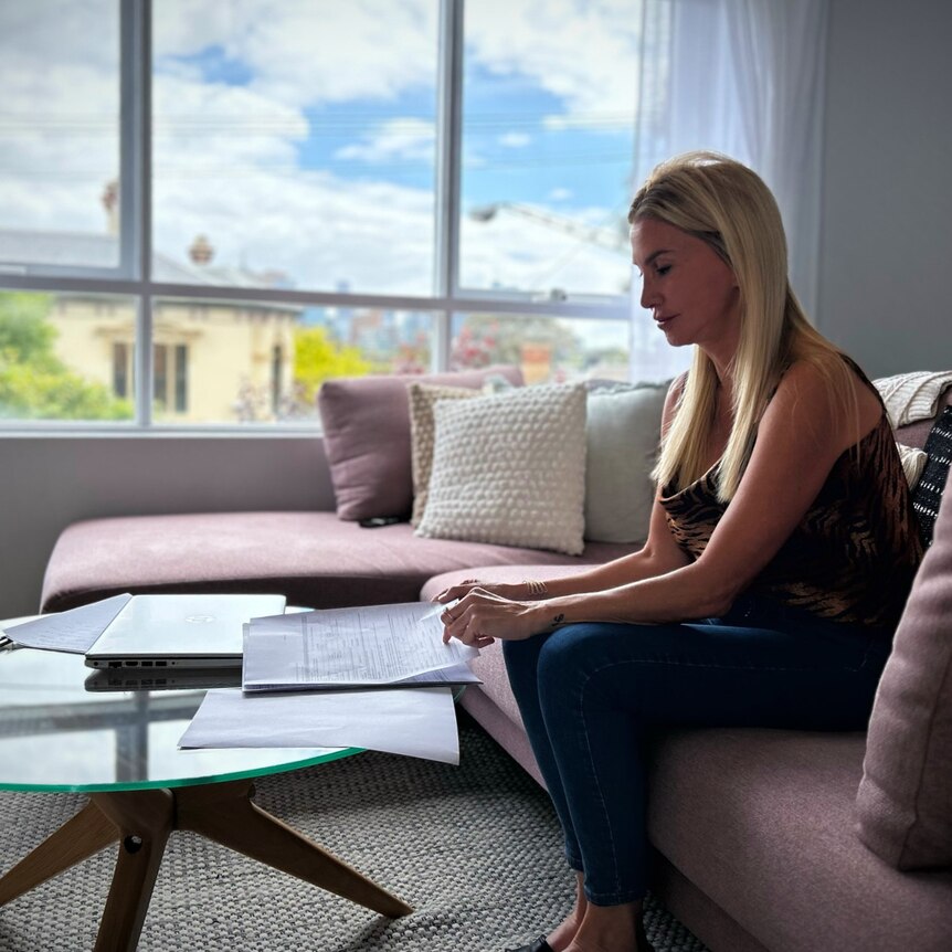 A woman with long blonde hair sits on a pink couch by a glass coffee table and looks at documents.