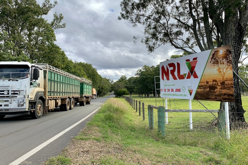 A cattle truck with a white cab and green trailer drives past the NRLX sign.