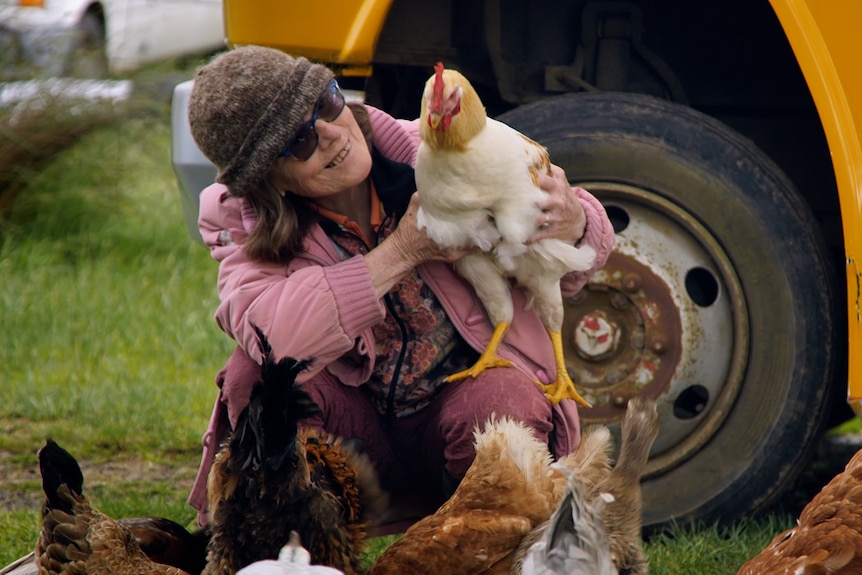 Woman wearing winter clothing with hat and sunglasses on holding a cockerel with multiple hens at her feet. 