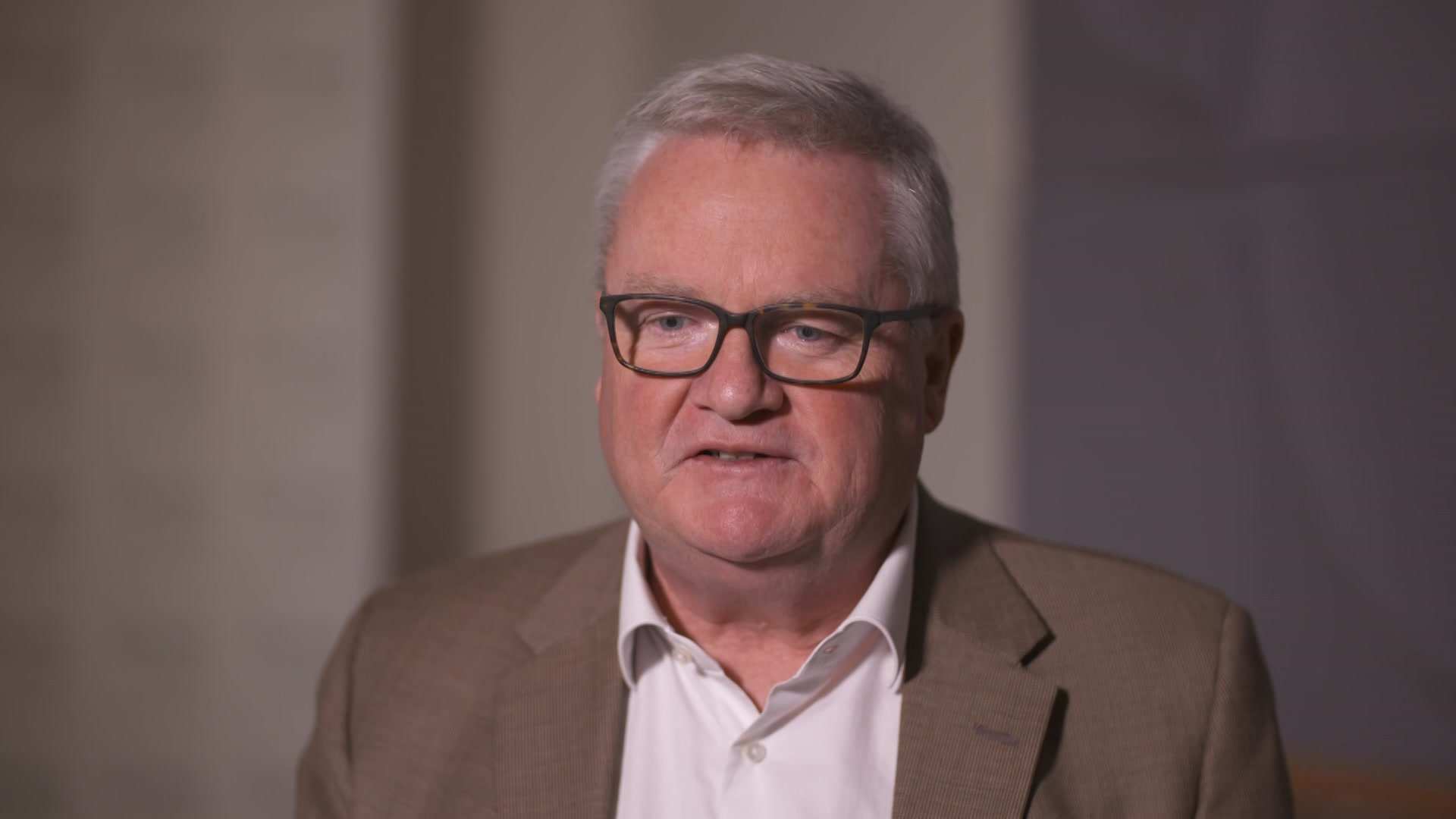 MCU of Peter Gordon sitting in a brown suit and open collared white shirt.