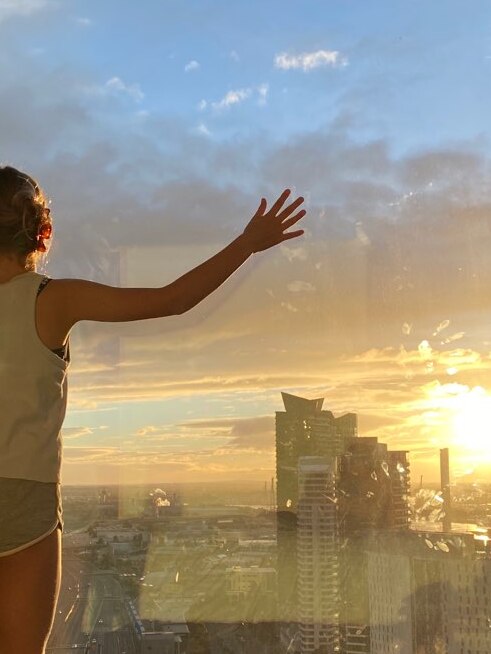 A young girl with her hands up against a big glass window in a high rise tower, looking over the cityscape