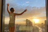 A young girl with her hands up against a big glass window in a high rise tower, looking over the cityscape