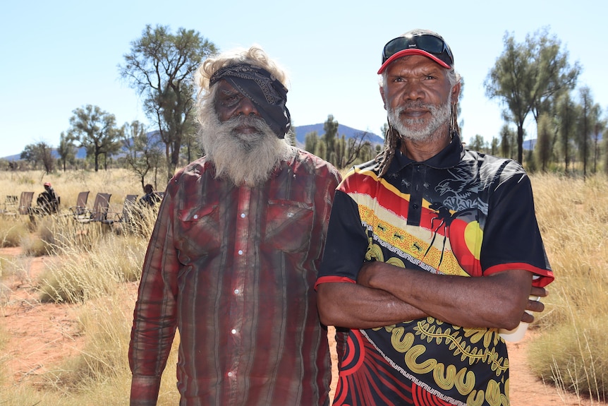 A man wearing black bandana over one eye, with grey hair and beard standing next to a younger man