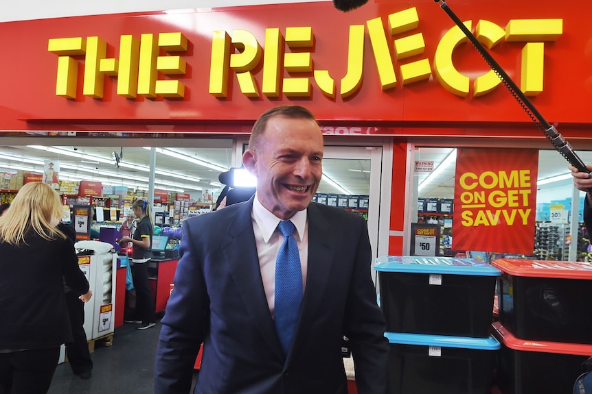 Tony Abbott smiles with the words "The Reject" behind him in a sign