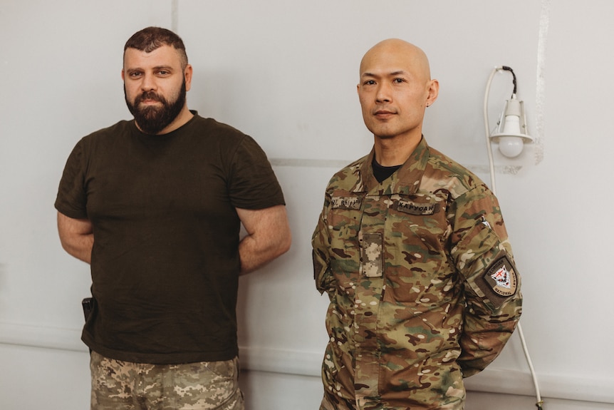 Two men in military gear stand side by side with their arms behind their backs in a white room