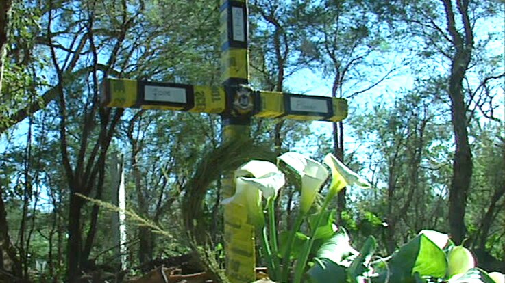 A cross wrapped in yellow police tape in a bushland setting with arum lilies in front of it.