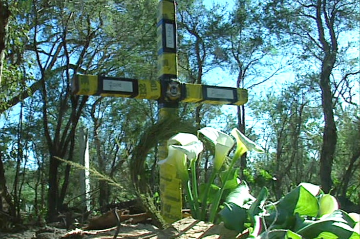 A cross wrapped in yellow police tape in a bushland setting with arum lilies in front of it.
