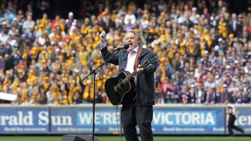 Mike Brady holds a guitar and sings in front of a large crowd at the MCG.
