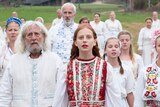 A group of people in white clothes covered in colourful flowers stare mournfully into the distance.